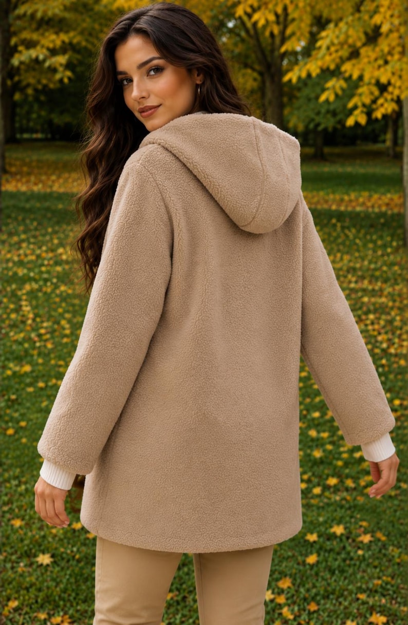 May include: A beige hooded jacket made of textured fleece material. The jacket features long sleeves with white ribbed cuffs and a hood. The design is simple and functional, providing a soft, fuzzy appearance with a consistent light brown color.