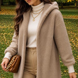 May include: A beige hooded teddy coat with a textured fleece finish, worn over a white ribbed turtleneck sweater and tan trousers, paired with a brown leather handbag featuring a chunky gold chain strap and a gold layered necklace.
