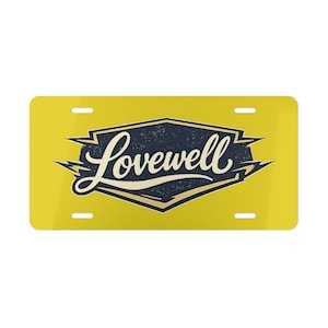 May include: A rectangular yellow number plate featuring a dark blue, shield-shaped emblem in the centre. The word Lovewell is written in a cream-coloured script font across the emblem, which is accented by sharp, lightning-like points on the sides.