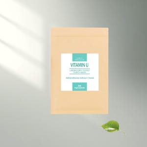 Könnte beinhalten: Ein beigefarbener Beutel mit einem weißen Etikett mit der Aufschrift "Vitamin U" und "Laboratory Tested Purity - 99.9%". Das Etikett enthält auch den Text "Methylmethionine Sulfonium Chloride" und "500 mg/Kapsel". Ein grünes Blatt befindet sich in der unteren rechten Ecke.