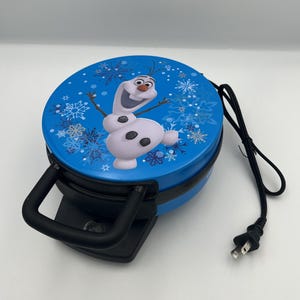 May include: A round blue waffle maker featuring a graphic of the character Olaf on the lid. The appliance has a black handle, a black base, and a black power cord with a standard two-prong electrical plug extending from the side of the unit.