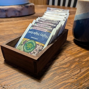 Peut inclure: Un présentoir en bois contenant des sachets de thé individuels. Les sachets portent l'inscription "Nighty Night Extra" avec du texte indiquant des ingrédients biologiques comme la mélisse et la valériane. Le présentoir est sur une table en bois, près d'une tasse bleue et blanche.