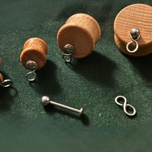 Puede incluir: Tapones para los oídos de madera de varios tamaños con detalles plateados. También hay una barra plateada y un pendiente en forma de ocho. Los artículos están sobre una superficie verde oscuro.