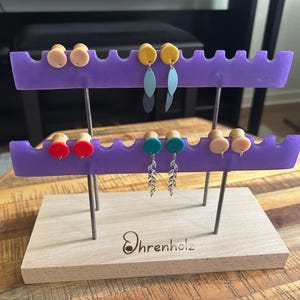 Könnte beinhalten: Ein Schmuckständer aus Holz mit zwei violetten Harzstangen mit gezackten Kanten. Der Ständer hält mehrere Paare Ohrplugs und Hängeohrringe in verschiedenen Farben, darunter Beige, Gelb, Rot, Türkis und Blau, mit dem Schriftzug Ohrenholz.