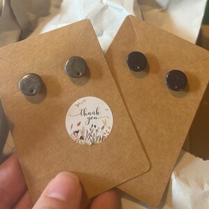 Könnte beinhalten: Zwei Paar runde Ohrstecker auf braunen Ohrringkarten. Ein Paar ist eine strukturierte silberne Farbe, und das andere ist dunkelbraun. Ein Aufkleber mit den Worten "Thank you" und floralen Akzenten ist sichtbar.