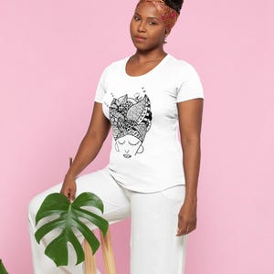 Może przedstawiać: Biały t-shirt z czarno-białą grafiką przedstawiającą twarz kobiety i chustę na głowie. Kobieta ma na sobie białe spodnie i wzorzystą chustę na głowie. Trzyma duży zielony liść. Tło jest r&oacute;żowe.
