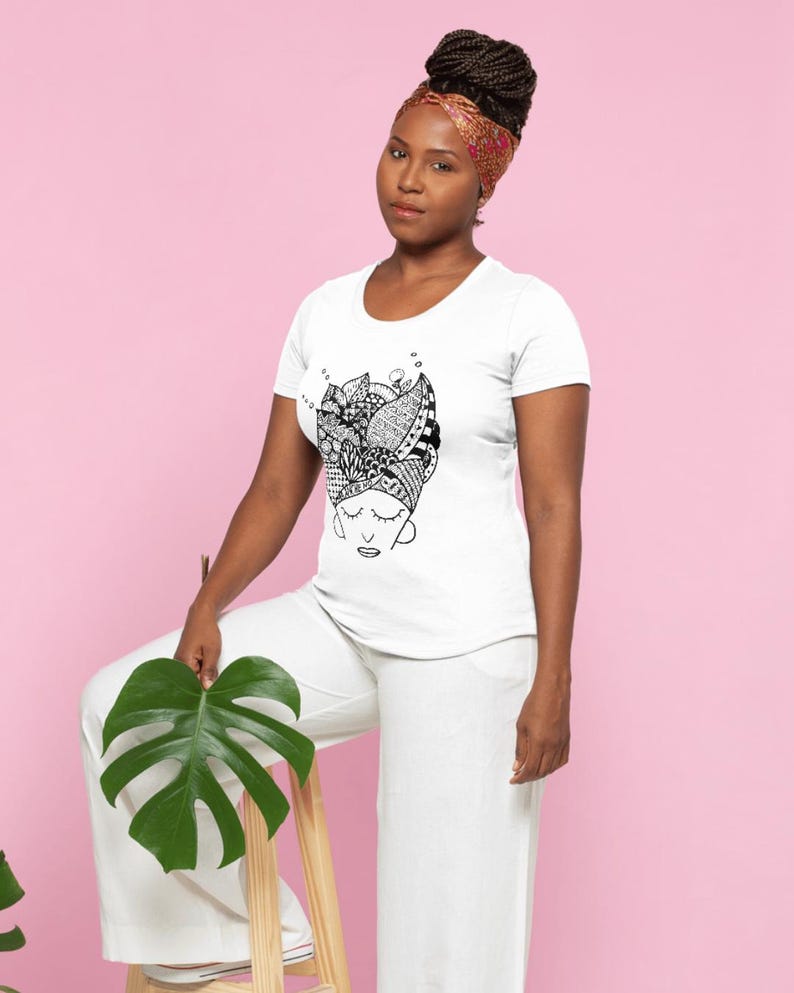 Może przedstawiać: Biały t-shirt z czarno-białą grafiką przedstawiającą twarz kobiety i chustę na głowie. Kobieta ma na sobie białe spodnie i wzorzystą chustę na głowie. Trzyma duży zielony liść. Tło jest r&oacute;żowe.