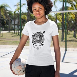 May include: White t-shirt featuring a black and white illustration of a person's head with a detailed design. The design includes closed eyes and intricate patterns. The person is holding a soccer ball.
