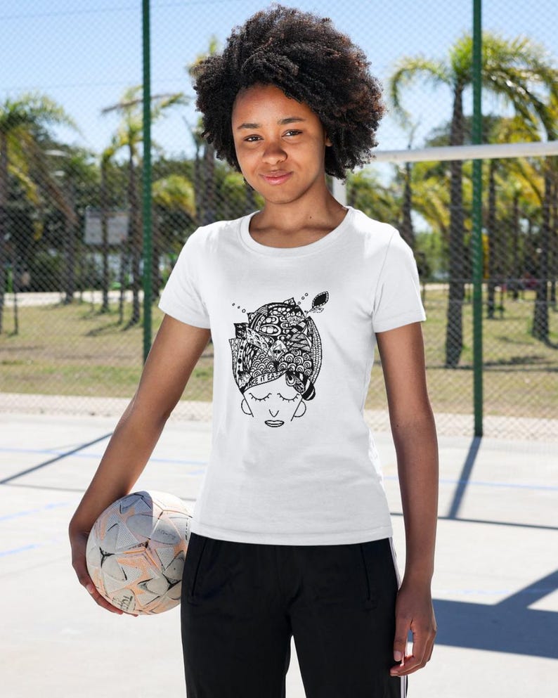May include: White t-shirt featuring a black and white illustration of a person's head with a detailed design. The design includes closed eyes and intricate patterns. The person is holding a soccer ball.