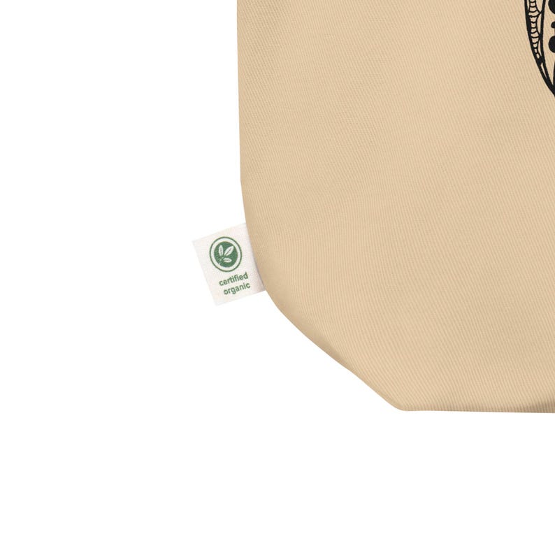 Peut inclure: Sac fourre-tout beige avec un motif noir. Une petite &eacute;tiquette blanche avec les mots "certified organic" et un logo de feuille verte est attach&eacute;e au sac. Le sac est fait d'une mati&egrave;re douce et naturelle.