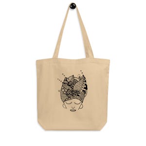 Pu&ograve; includere: Borsa tote beige con un'illustrazione in bianco e nero di un volto con gli occhi chiusi e un copricapo a motivi. La borsa ha manici lunghi e un design semplice, adatta per trasportare vari oggetti.