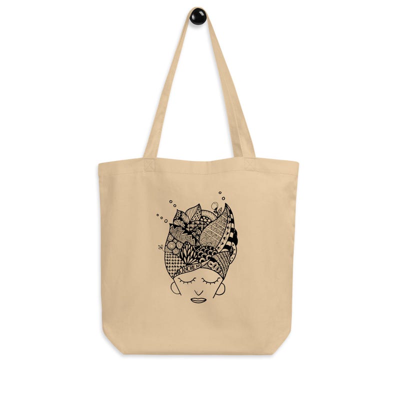 Pu&ograve; includere: Borsa tote beige con un'illustrazione in bianco e nero di un volto con gli occhi chiusi e un copricapo a motivi. La borsa ha manici lunghi e un design semplice, adatta per trasportare vari oggetti.