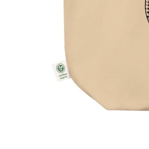Pu&ograve; includere: Una borsa tote beige con un'etichetta "certificato biologico". L'etichetta &egrave; bianca con un disegno a forma di foglia verde. La borsa &egrave; realizzata in un materiale morbido e naturale.