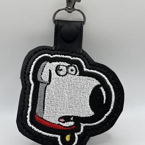 May include: Embroidered keychain featuring a cartoon dog character with a white face, black nose, and red collar. The keychain has a black border and a metal clasp for attaching to keys or bags.