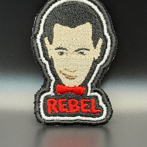 May include: Embroidered patch featuring a stylized portrait with black hair, a tan face, and a red bow tie. The word "REBEL" is embroidered in red letters at the bottom, outlined in white. The patch has a black border.