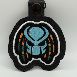 May include: Black leather keychain featuring an embroidered Predator design. The design includes a blue face with orange, yellow, and green accents, outlined in white. The keychain has a black snap closure.