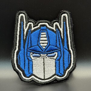 May include: Embroidered patch featuring the face of Optimus Prime in blue, white, and black. The patch has a detailed design with a black border and is likely for application on clothing or accessories.