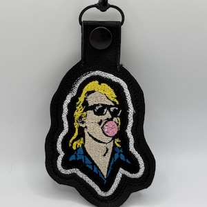 May include: Black embroidered keychain featuring a cartoon image of a person with sunglasses blowing a bubblegum bubble. The person has blonde hair and is wearing a blue shirt. The keychain has a black leather strap and a metal clasp.