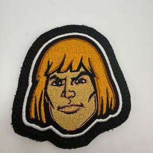 May include: Embroidered patch featuring a cartoon image of He-Man with yellow hair and a stern expression. The patch has a black border with a white outline, and the character's face is tan with pink accents.