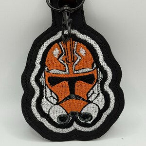 May include: Black keychain with an embroidered orange and white helmet design. The helmet has a detailed, stylized appearance, with a black border and a metal clasp for attachment. The design is reminiscent of a popular science fiction theme.
