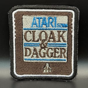 May include: Embroidered patch featuring the text "ATARI CLOAK & DAGGER" in blue and brown. The patch has a brown border with a white outline and a black edge. The Atari logo is at the bottom.