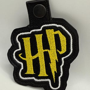 May include: Black and yellow embroidered patch featuring the letters "HP" with a lightning bolt design. The patch has a white border and a black leather backing with a snap closure.