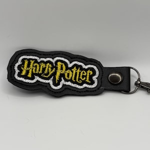May include: Black keychain with the embroidered text "Harry Potter" in yellow and white. The keychain has a black leather strap and a silver-colored clasp.