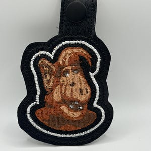 May include: Black and brown embroidered keychain featuring a cartoon character with a black border and white stitching. The keychain has a black leather strap with a snap closure.