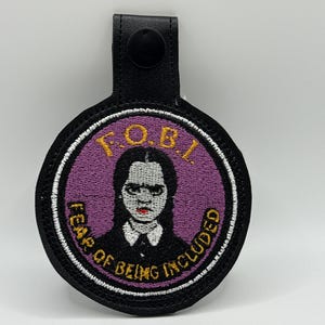 May include: A black and purple patch featuring a portrait of a character with the text "F.O.B.I. Fear of Being Included". The patch has a black leather strap with a black snap closure.