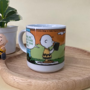 May include: A white ceramic mug featuring a Peanuts comic strip illustration of Charlie Brown. The mug has a handle and is set on a wooden tray with a small plant and a Charlie Brown figurine.