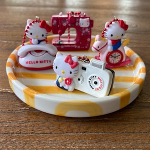 Puede incluir: Una colección de miniaturas con temática de Hello Kitty en un plato a rayas. Las miniaturas incluyen un teléfono, una máquina de coser, una cámara y un caballito balancín, todos con el personaje icónico. El plato tiene rayas amarillas y blancas.