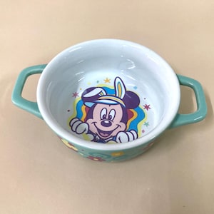 Può includere: Una piccola ciotola rotonda in ceramica con due manici. La ciotola è azzurra con una grafica di Topolino al centro. La grafica mostra Topolino con un cappello e sorridente. La ciotola ha un motivo floreale sul bordo.
