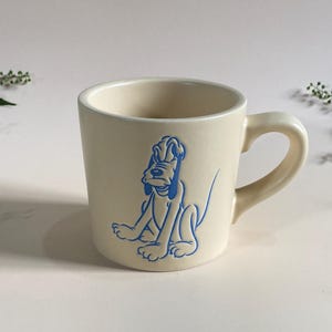 Peut inclure: Mug en céramique crème avec une illustration d'un chien de dessin animé bleu. La tasse a une anse arrondie et une forme droite. Le chien est assis. La tasse est sur une surface blanche.