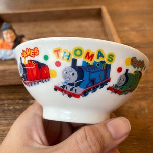 Puede incluir: Cuenco de cerámica blanca con ilustraciones de Thomas el Tren y sus amigos. El cuenco tiene la palabra "THOMAS" en amarillo e imágenes de James, Thomas y Percy. Un cuenco colorido para niños.