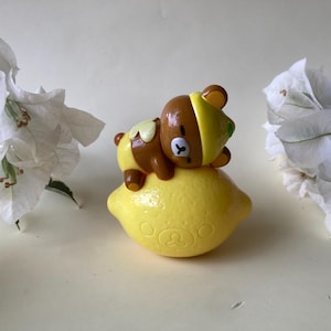 May include: A brown and yellow bear figurine wearing a lemon hat, resting on a yellow lemon. The lemon has a bear face design on it. White flowers are in the background.