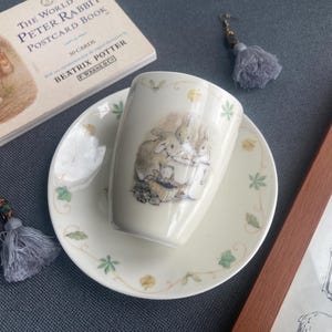 Puede incluir: Un juego de taza y plato de cerámica color crema con una ilustración impresa de tres conejos. La taza y el plato están decorados con un borde delicado de pequeñas hojas verdes y flores amarillas. La taza muestra conejos cerca de una cesta.