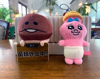 May include: Two plush toys: a brown mushroom with a smiling face and a pink bunny wearing a yellow hat and blue bow. The mushroom holds a sign with Japanese text. The bunny wears a diaper.