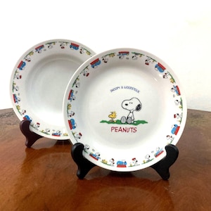 May include: Two white ceramic plates featuring Snoopy and Woodstock. The front plate displays the text "SNOOPY & WOODSTOCK" and "PEANUTS" with a central illustration of the characters. Both plates have a repeating border pattern of the characters.