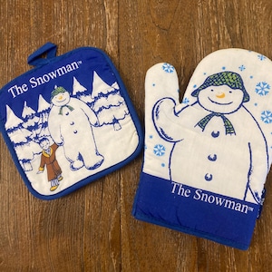 Puede incluir: Un juego de agarradera y guante de horno azul y blanco con un diseño de muñeco de nieve. La agarradera es cuadrada con un lazo, y el guante de horno tiene forma de mano. Ambos artículos tienen el texto "The Snowman".