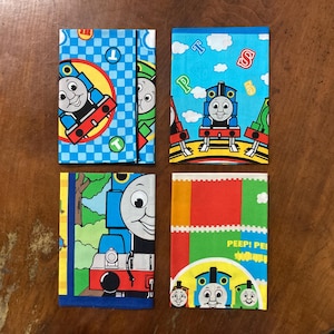 Könnte beinhalten: Vier farbenfrohe Stoffartikel mit der Zeichentrickfigur Thomas, die kleine Lokomotive. Die Artikel haben blaue, rote, grüne und gelbe Hintergründe mit Bildern von Thomas. Ein Artikel hat den Text "PEEP! PEEP!"