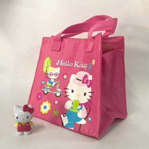 Pode incluir: Uma sacola rosa Hello Kitty com alças combinando e uma pequena estatueta Hello Kitty. A sacola apresenta ilustrações de Hello Kitty e o texto "Hello Kitty". A estatueta é branca com um laço vermelho e um vestido amarelo.