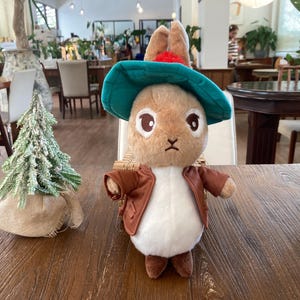 May include: A plush rabbit toy wearing a green hat and brown jacket, with a small Christmas tree in the background. The rabbit has large, expressive eyes and is posed on a wooden table. The tree is in a burlap sack.