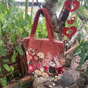 以下が含まれることがあります： 花柄とキャラクター刺繍が施された赤いトートバッグ。バッグは長方形で、大きく開口部があり、2つの長いハンドルが付いています。デザインは、さまざまなカラフルな花と漫画のキャラクターが特徴です。バッグは緑の近くの屋外に展示されています。