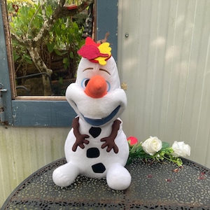 May include: A plush Olaf doll from the movie Frozen, featuring a white body, black buttons, and a carrot nose. The doll has a blue collar, brown stick arms, and a colorful headpiece. White roses and red rosebuds are next to the doll.