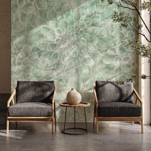 May include: Two gray upholstered armchairs with wooden frames are positioned in front of a wall with a large, faded green floral pattern. A small, round wooden table with a black metal base and a beige vase sit between the chairs.