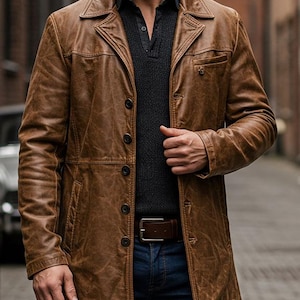 May include: A brown leather coat with a button-up front, a notched collar, and a chest pocket. The coat is worn over a dark gray sweater and blue jeans. A brown leather belt is visible.