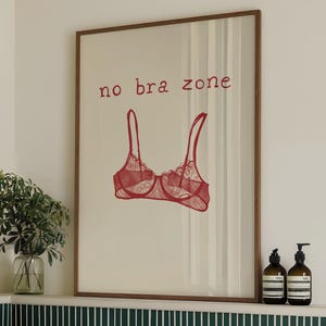 May include: A framed print with a red bra illustration and the text "no bra zone." The artwork is displayed above a white bathtub with a green towel and green and white tile. Two brown bottles with black pumps are on the shelf.