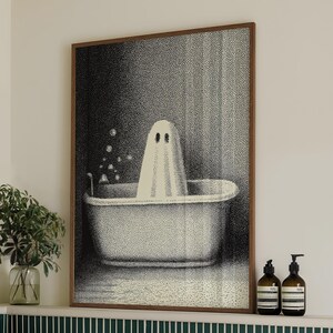 May include: A framed black and white print of a ghost in a bathtub. The bathroom setting includes a white bathtub, green and white tile, and a shelf with soap bottles and a plant. A stack of white towels with green trim is on the tub.