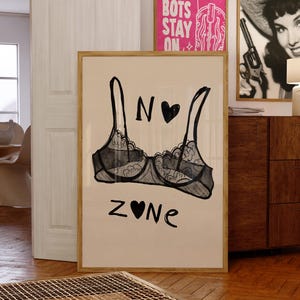 May include: A framed art print featuring a black and white illustration of a bra with the words "N ♥ Zone". The artwork is displayed in a room with other framed art and wooden furniture.