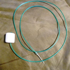 May include: A green, circular, flexible wire with a white, rectangular foam piece attached to one end and a silver needle-like object at the other. The wire is lying on a textured, olive-green surface.